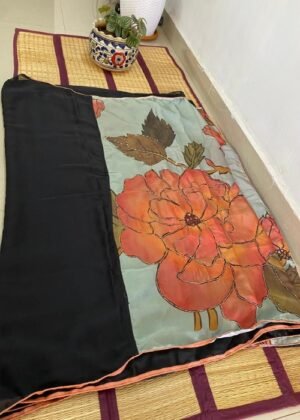 Black saree with a large floral hand-painted design displayed on a woven mat with decorative potted plants.